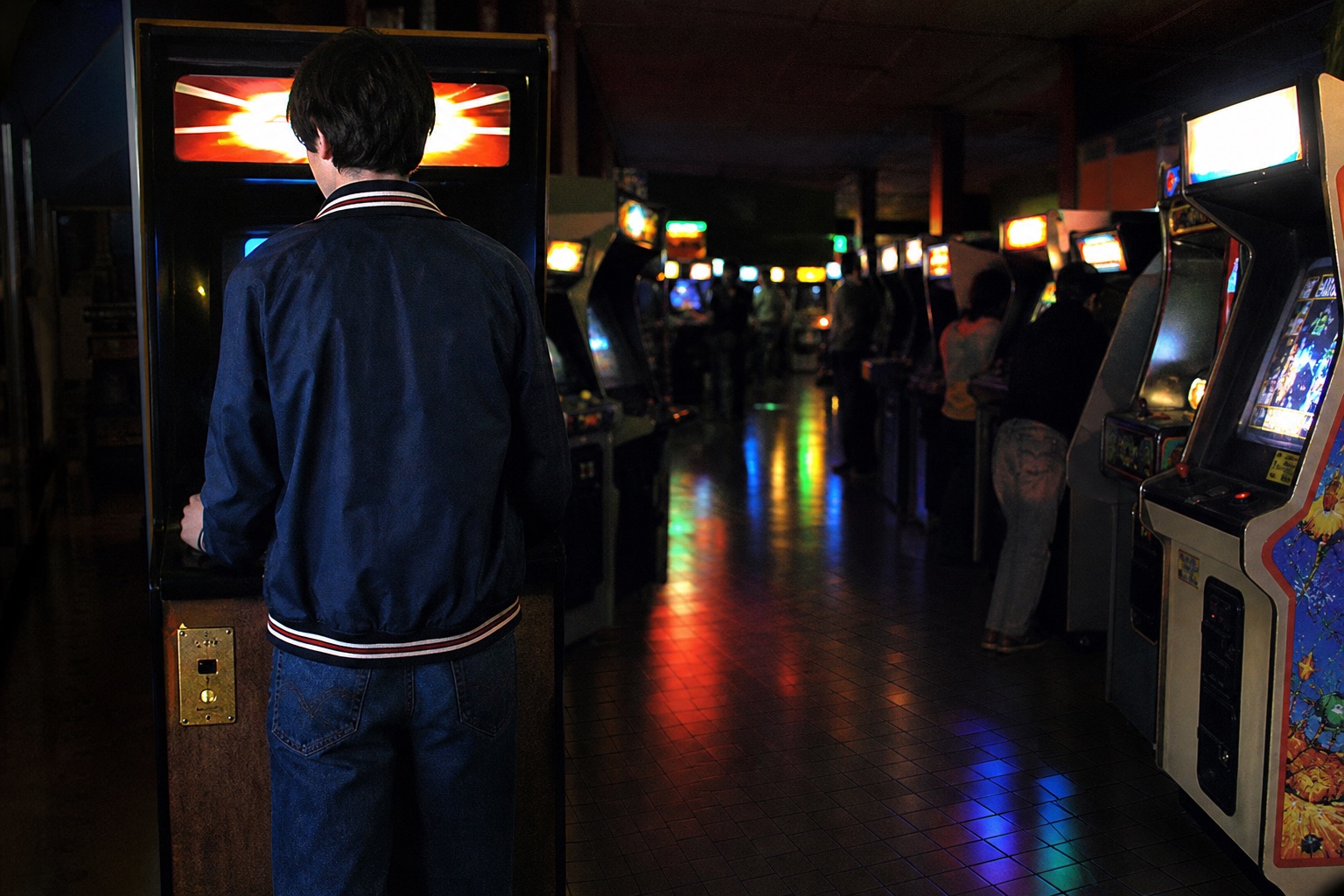 Salle d'arcade française de 1984, rangées de bornes allumées dans la pénombre, reflets colorés sur le carrelage